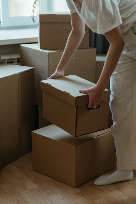 A young woman with long dark hair sitting cross-legged on a wooden floor surrounded by numerous medium-sized brown cardboard boxes, some with red 'Fragile' labels. She is inside a room with a textured